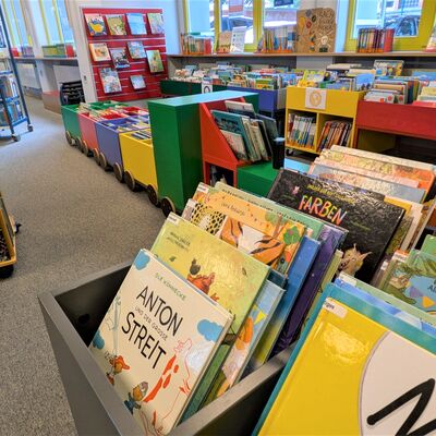 Bild vergr&ouml;&szlig;ern: Blick in eine farbenfrohe Kinderbibliothek mit zahlreichen Bilderb&uuml;chern in niedrigen Regalen und bunten B&uuml;cherkisten, die wie ein kleiner Zug gestaltet sind. Im Vordergrund ist ein Buch mit dem Titel &raquo;Anton und der gro&szlig;e Streit&laquo; in einer grauen Kiste deutlich zu erkennen.