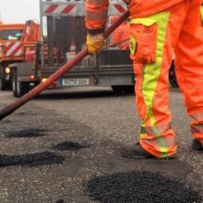 Ein Bauarbeiter in oranger Warnkleidung arbeitet an einer Stra&szlig;e. Er bessert L&ouml;cher im Asphalt aus. Im Hintergrund steht ein orangefarbenes Baufahrzeug - Dies ist ein Button, der zu den weiterf&uuml;hrenden Texten zur Ausbildung zum Stra&szlig;enw&auml;rter f&uuml;hrt.
