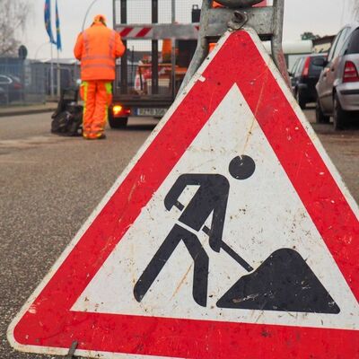 Bild vergr&ouml;&szlig;ern: Ein dreieckiges Warnschild weist auf eine Baustelle hin. Es steht gro&szlig; im Vordergrund. Im unscharfen Hintergrund ist ein Bauarbeiter in orangefarbener Kleidung an einer Stra&szlig;e zu sehen - Dies ist ein Button, der zu den weiterf&uuml;hrenden Texten zur Ausbildung zum Stra&szlig;enw&auml;rter f&uuml;hrt.