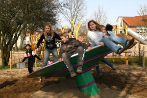 Das Foto zeigt eine Gruppe von sechs Kindern und Jugendlichen, die an einem sonnigen Tag sichtlich Spa&szlig; auf einem Spielplatz haben. Im Mittelpunkt steht ein gro&szlig;es, rotierendes Spielger&auml;t - eine schr&auml;g gestellte Drehscheibe (oft auch als &raquo;Supernova&laquo; oder rotierende Wippe bezeichnet). Das Ger&auml;t steht auf einem dicken, gr&uuml;nen Mittelfu&szlig;, der im sandigen Boden verankert ist. Die Scheibe selbst ist metallisch mit gr&uuml;nen und lila Akzenten. Durch die Neigung und Drehung ist die Szene voller Dynamik.