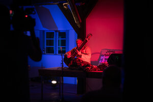 Bild vergr&ouml;&szlig;ern: Eine Weitwinkelaufnahme eines Sitarspielers auf einer kleinen B&uuml;hne im Innenbereich. Die Szene wird von dramatischem blauem und rotem Licht beleuchtet, wobei das blaue Licht von links und das rote Licht von rechts kommt. Der Sitarspieler sitzt auf einem Teppich in der Mitte der B&uuml;hne und tr&auml;gt eine traditionelle helle Tunika.