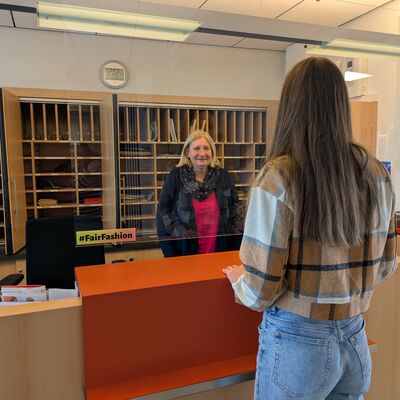 Bild vergr&ouml;&szlig;ern: Eine junge Frau mit langen Haaren steht an einem h&ouml;lzernen Empfangstresen und l&auml;sst sich von einer l&auml;chelnden Mitarbeiterin beraten. Die Mitarbeiterin befindet sich hinter einer gl&auml;sernen Trennscheibe, im Hintergrund ist ein gro&szlig;es Aktenregal (Postf&auml;cher) zu sehen. Auf dem Tresen klebt ein kleiner Aufkleber mit dem Hashtag &raquo;Fairfashion&laquo;.