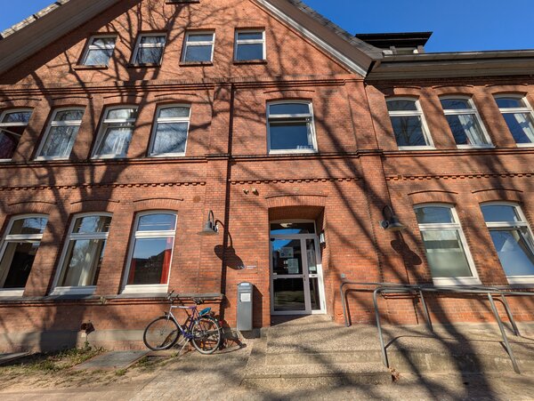 Die Au&szlig;enansicht eines mehrst&ouml;ckigen Backsteingeb&auml;udes bei sonnigem Wetter. Im Zentrum f&uuml;hrt eine kleine Steintreppe mit Metallgel&auml;nder zu einer gl&auml;sernen Eingangst&uuml;r. Links neben dem Eingang steht ein Fahrrad an der Hauswand. Lange Schatten von B&auml;umen fallen &uuml;ber die gesamte Fassade und die vielen Fenster des Geb&auml;udes.
