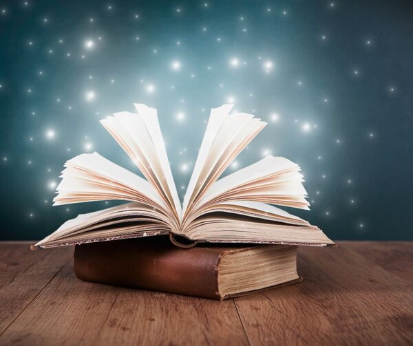 Ein aufgeschlagenes, antikes Buch liegt auf einem geschlossenen Buch mit braunem Ledereinband auf einem Holztisch. Die Seiten des oberen Buches f&auml;chern sich nach oben auf. Vor einem dunkelblauen Hintergrund steigen helle, funkelnde Lichtpunkte und Glitzereffekte aus dem Buch empor, die eine magische, m&auml;rchenhafte Atmosph&auml;re erzeugen.