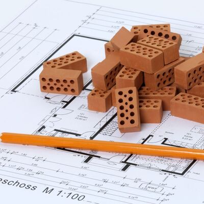 Bild vergr&ouml;&szlig;ern: Ein Symbolbild zum Thema Architektur und Bauwesen. Auf einem detaillierten Grundrissplan f&uuml;r ein Erdgeschoss im Ma&szlig;stab 1:100 liegt ein kleiner Haufen Miniatur-Ziegelsteine aus r&ouml;tlichem Ton. Im Vordergrund liegt ein spitzer, orangefarbener Bleistift quer &uuml;ber dem Plan.