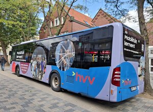 Die R&uuml;ck- und Seitenansicht eines hvv-Busses, der als mobiler Lernort gestaltet ist. Das Fahrzeug ist hellblau und fliederfarben foliert und zeigt kreisf&ouml;rmige Fotos von lernenden Jugendlichen. Auf dem Heck und an der Seite steht in fetten wei&szlig;en Buchstaben der Slogan: &raquo;Mobilit&auml;t lernen! Mit dem hvv Schulprojekt&laquo;. Der Bus parkt in einer gepflasterten Fu&szlig;g&auml;ngerzone vor einem Backsteingeb&auml;ude und B&auml;umen.