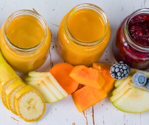Eine Draufsicht auf drei offene Gl&auml;ser mit buntem Babybrei (zwei in Gelb-Orange, eines in Dunkelrot) auf einem wei&szlig;en Holztisch. Davor liegen frisch aufgeschnittene Obst- und Gem&uuml;sest&uuml;cke: Bananenscheiben, Birnenspalten, orangefarbene K&uuml;rbisw&uuml;rfel sowie eine Brombeere und eine Heidelbeere.