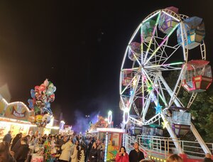 Eine n&auml;chtliche Aufnahme eines belebten Jahrmarkts. Im rechten Vordergrund ragt ein gro&szlig;es, hell erleuchtetes Riesenrad mit wei&szlig;en Speichen und bunten Gondeln in den dunklen Nachthimmel. Links im Bild sieht man ein gro&szlig;es B&uuml;ndel bunter Luftballons mit verschiedenen Motiven. Zwischen dem Riesenrad und den beleuchteten Buden dr&auml;ngen sich zahlreiche Besucher in einer Gasse, &uuml;ber der stellenweise leichter Nebel oder Rauch schwebt.