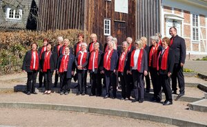 Ein Gruppenfoto eines Frauenchors im Freien. Etwa 20 S&auml;ngerinnen stehen in zwei Reihen vor dem rustikalen Geb&auml;ude der Stankt Salvatoris Kirche. Alle Chormitglieder tragen eine einheitliche Garderobe bestehend aus schwarzen Hosen, dunklen Blusen oder Sakkos und auff&auml;lligen, langen roten Schals. Die Frauen l&auml;cheln und wirken entspannt; im Hintergrund sind kahle Str&auml;ucher und helles Sonnenlicht zu sehen.