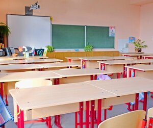 Bild vergr&ouml;&szlig;ern: Modernes Klassenzimmer: Ein heller Unterrichtsraum mit mehreren Reihen leerer Sch&uuml;lertische und Holzst&uuml;hlen mit roten Gestellen. An der Stirnseite befinden sich eine gro&szlig;e gr&uuml;ne Schultafel sowie ein digitales Whiteboard mit einem dar&uuml;ber installierten Beamer.