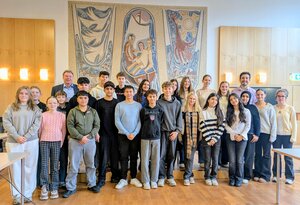 Bild vergr&ouml;&szlig;ern: Gruppenfoto der ANS-Sch&uuml;ler: Eine gro&szlig;e Gruppe von etwa 20 Sch&uuml;lerinnen und Sch&uuml;lern der Alfred-Nobel-Schule (ANS) posiert f&uuml;r ein gemeinsames Foto im Sitzungssaal des Rathauses. Sie stehen vor drei gro&szlig;en, k&uuml;nstlerisch gestalteten Wandteppichen, die verschiedene Motive in Blau-, Rot- und Goldt&ouml;nen zeigen. Begleitet wird die Gruppe von zwei erwachsenen M&auml;nnern, die links und rechts au&szlig;en stehen.