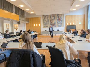 Bild vergr&ouml;&szlig;ern: Sch&uuml;lerbesuch im Rathaus: Eine Gruppe von Sch&uuml;lerinnen und Sch&uuml;lern der Alfred-Nobel-Schule (ANS) sitzt an einer u-f&ouml;rmigen Tischreihe im hellen Sitzungssaal des Geesthachter Rathauses. Ein Mann steht im Zentrum des Raumes und spricht zur Gruppe; im Hintergrund h&auml;ngen drei gro&szlig;e, vertikale Wandteppiche mit maritimen und abstrakten Motiven.