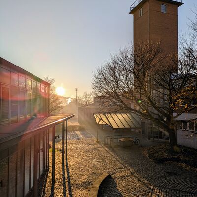Bild vergr&ouml;&szlig;ern: Ein stimmungsvolles Foto des Geesthachter Rathausr&uuml;ckplatzes bei Sonnenaufgang. Die tiefstehende Sonne wirft lange Schatten auf das Kopfsteinpflaster und l&auml;sst das moderne Verwaltungsgeb&auml;ude sowie den markanten Backsteinturm der benachbarten Feuerwehr in warmem, goldenem Licht erstrahlen.