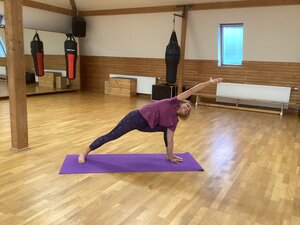 Eine Frau praktiziert Yoga in einem hellen Trainingsraum mit Holzfu&szlig;boden. Sie befindet sich in der Position des gestreckten Seitenwinkels auf einer lila Matte, wobei ein Arm weit &uuml;ber den Kopf nach vorne gestreckt ist.