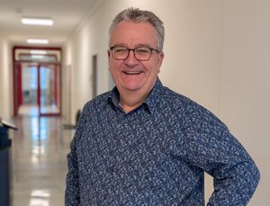 Bild vergr&ouml;&szlig;ern: Heiner Ro&szlig;mann steht in einem hellen Flur mit wei&szlig;en W&auml;nden und gl&auml;nzendem Boden. Er tr&auml;gt ein dunkelblaues Hemd mit feinem Muster und hat eine Hand in die Hosentasche gesteckt. Im Hintergrund sind T&uuml;ren und ein Fenster zu sehen, die Tageslicht hereinlassen.