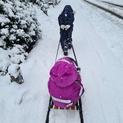 Bild vergr&ouml;&szlig;ern: Symbolbild zum Winter - Zwei Personen auf einem schneebedeckten Weg, eine zieht einen Schlitten mit der anderen Person sitzend darauf, umgeben von schneebedeckten Stra&szlig;en.