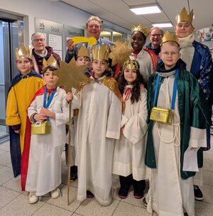 Bild vergr&ouml;&szlig;ern: Mehrere Kinder stehen in einem Flur des Geesthachter Rathauses. Sie tragen festliche Gew&auml;nder in Wei&szlig;, Blau, Rot und Gr&uuml;n sowie goldene Kronen auf dem Kopf. Einige halten gro&szlig;e goldene Sterne, andere tragen Umh&auml;nge mit glitzernden Kragen. Um den Hals h&auml;ngen goldene Sammelb&uuml;chsen. Im Hintergrund sind Wandtafeln mit dem Schriftzug 'Stadt Geesthacht' und weitere Personen zu sehen. Die Szene zeigt den Besuch der Sternsinger am 6. Januar 2026, bei dem sie B&uuml;rgermeister Olaf Schulze einen Segen f&uuml;r die Stadt &uuml;berbringen.