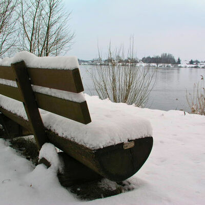 Bild vergr&ouml;&szlig;ern: Eine mit Schnee bedeckte Bank in einer winterlichen Landschaft.