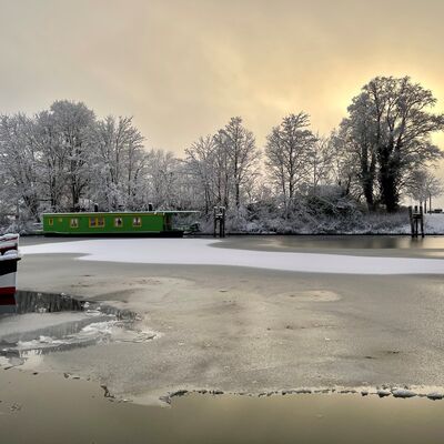 Bild vergr&ouml;&szlig;ern: Vereister Hafen mit gr&uuml;nem Hausboot am Ufer und schneebedeckten B&auml;umen im Hintergrund