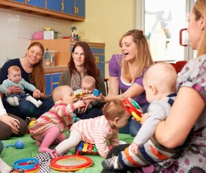 Eine Gruppe von Frauen mit Babys auf dem Scho&szlig; sitzt in einem Spielzimmer und mehrere Babys krabbeln einer Decke. Symbolbild zum Thema Babytreff.