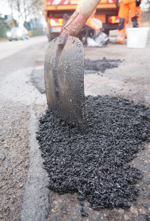 Bild vergr&ouml;&szlig;ern: Eine Person verteilt mit einer Schaufel Asphalt, w&auml;hrend Mitarbeitende der St&auml;dtischen Betriebe Schlagl&ouml;cher reparieren.