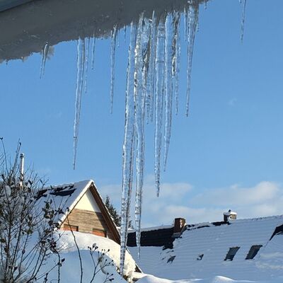 Bild vergr&ouml;&szlig;ern: Symbolbild zum Winter - Mehrere lange Eiszapfen h&auml;ngen von einem Dachvorsprung &uuml;ber schneebedeckten Hausd&auml;chern unter blauem Himmel