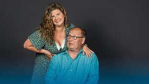 Eine Frau mit lockigem Haar in einem gemusterten Kleid steht hinter einem sitzenden Mann in einem karierten Hemd und legt eine Hand auf seine Schulter vor dunklem Hintergrund.