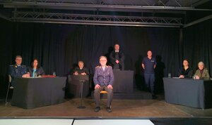 Acht Personen auf einer B&uuml;hne mit schwarzem Vorhang, eine sitzt vorne auf einem Stuhl in Uniform, zwei stehen hinter einem Tisch, vier sitzen an einem weiteren Tisch mit Mikrofon.