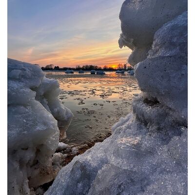 Nahaufnahme von vereisten Eisberg am Elbufer mit Blick auf eine teilweise zugefrorene Wasserfl&auml;che und einen Sonnenuntergang im Hintergrund