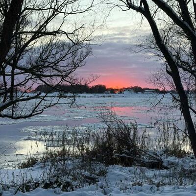 Bild vergr&ouml;&szlig;ern: Schneebedeckte Landschaft mit einem Sonnenuntergang hinter den B&auml;umen, der den Himmel in warmen Farben erleuchtet.