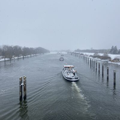 Bild vergr&ouml;&szlig;ern: Verschneite Uferlandschaft mit kahlen B&auml;umen und einem Frachtschiff auf ruhigem Fluss im Winter