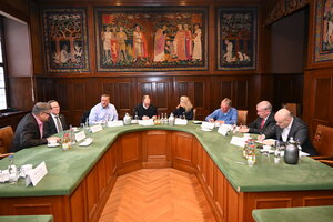 Bild vergr&ouml;&szlig;ern: Eine Gruppe von sieben Personen sitzt an einem gro&szlig;en, hufeisenf&ouml;rmigen Tisch mit gr&uuml;ner Oberfl&auml;che in einem historischen Sitzungssaal. Die W&auml;nde sind mit dunklem Holz get&auml;felt und mit gro&szlig;formatigen, farbenfrohen Wandteppichen geschm&uuml;ckt, die mittelalterliche Szenen darstellen. Die Teilnehmenden, M&auml;nner und Frauen in gesch&auml;ftlicher Kleidung, sind in ein Gespr&auml;ch vertieft oder machen sich Notizen. Auf dem Tisch stehen Namensschilder, Getr&auml;nke und Kaffeegeschirr.