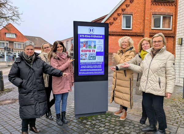 Sechs Frauen stehen im Freien auf einem gepflasterten Platz vor einem modernen digitalen Informations-Display. Sie l&auml;cheln und zeigen gemeinsam mit ausgestreckten H&auml;nden auf den Bildschirm. Das Display bewirbt ein &raquo;KTS-Kino-Fr&uuml;hst&uuml;ck&laquo; mit dem Film &raquo;COPA 71&laquo;. Im Hintergrund ist ein r&ouml;tliches Backsteingeb&auml;ude unter grauem Himmel zu sehen.