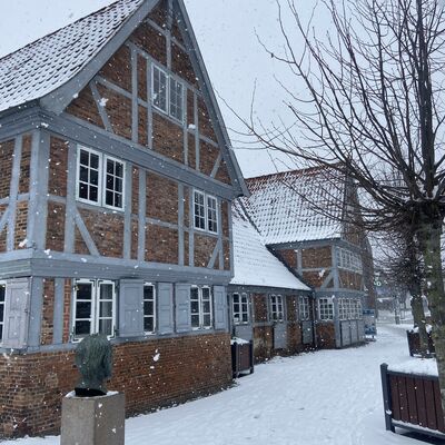 Bild vergr&ouml;&szlig;ern: Eine winterliche Au&szlig;enansicht des GeesthachtMuseums, bestehend aus zwei rechtwinklig zueinander stehenden Fachwerkgeb&auml;uden aus rotem Backstein und grauen Holzbalken. Im Vordergrund steht die Bronzeb&uuml;ste von Alfred Nobel auf ihrem Steinsockel, von hinten gesehen. Ein schneebedeckter Weg mit vielen Fu&szlig;spuren f&uuml;hrt zwischen den Geb&auml;uden hindurch, w&auml;hrend es leise weiterschneit.