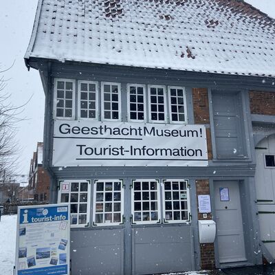 Bild vergr&ouml;&szlig;ern: Ein zweist&ouml;ckiges, historisches Geb&auml;ude mit grauer Fassade und schneebedecktem Ziegeldach w&auml;hrend eines leichten Schneefalls. Ein gro&szlig;es wei&szlig;es Banner zwischen den Fensterreihen tr&auml;gt die Aufschrift &raquo;GeesthachtMuseum! Tourist-Information&laquo;. Vor dem Geb&auml;ude steht links ein blauer Informationsst&auml;nder mit der Aufschrift &raquo;tourist-info&laquo;. Der Boden ist mit einer d&uuml;nnen Schneeschicht bedeckt.