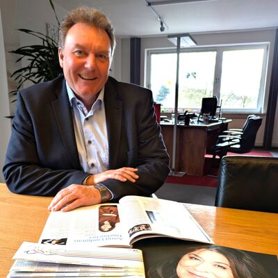 Ein l&auml;chelnder Mann (Olaf Schulze) in einem dunklen Sakko sitzt an einem Holztisch in einem B&uuml;ro. Vor ihm auf dem Tisch liegen verschiedene Werbematerialien f&uuml;r das Schleswig-Holstein Musik Festival (SHMF) 2026:
Vordergrund: Ein gro&szlig;es Plakat f&uuml;r die Veranstaltung &raquo;Astrid Lindgrens Tageb&uuml;cher&laquo; am 11. Juli in Geesthacht, auf dem das Portr&auml;t von Eva Mattes zu sehen ist.
Mittelfeld: Ein aufgeschlagenes Programmheft, das ebenfalls die Seite zu Astrid Lindgrens Tageb&uuml;chern zeigt, sowie ein Stapel gefalteter Flyer mit verschiedenen Ortsnamen (u. a. Ahrensburg, Brunsb&uuml;ttel, Hamburg, Kiel).
Hintergrund: Ein helles B&uuml;ro mit einem Schreibtisch am Fenster und einer Zimmerpflanze.
