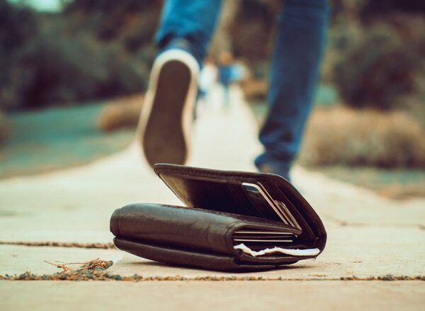 Unscharf im Hintergrund geht eine Person einen Weg entlang. Im Vordergrund liegt ein ge&ouml;ffnetes, dunkles Portemonnaie auf dem Boden, aus dem Karten und Papiere sichtbar herausragen. Die Person scheint den Verlust nicht zu bemerken und entfernt sich davon.