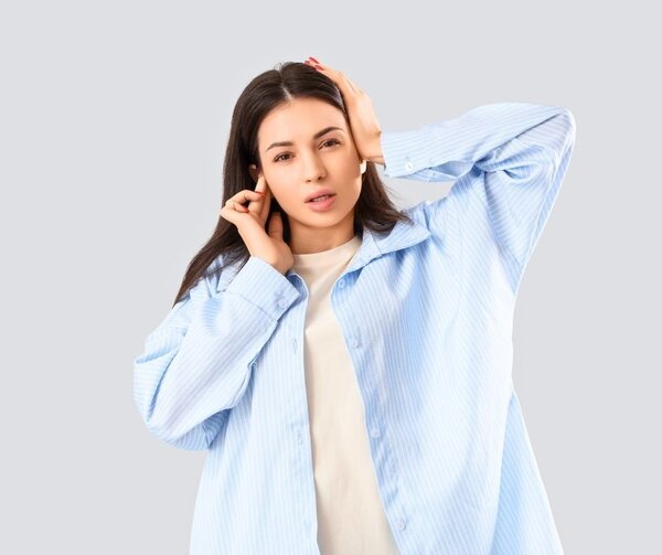 Eine junge Frau mit dunklen Haaren blickt mit einem genervten oder schmerzverzerrten Gesichtsausdruck direkt in die Kamera. Sie tr&auml;gt ein hellblau gestreiftes Hemd &uuml;ber einem beigen T-Shirt und h&auml;lt sich mit beiden H&auml;nden die Ohren zu, um sich gegen L&auml;rm zu sch&uuml;tzen. Der Hintergrund ist neutral und hellgrau.
