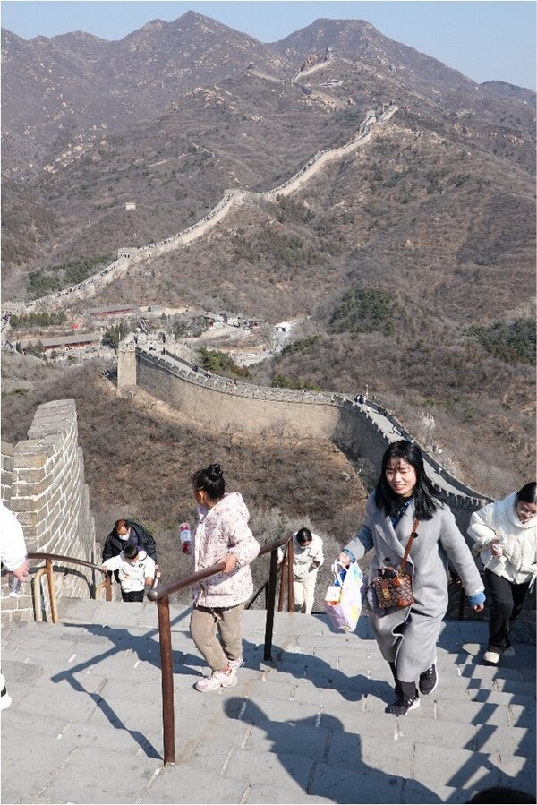 Bild vergr&ouml;&szlig;ern: Ein Blick von oben auf einen steilen Treppenabschnitt der Chinesischen Mauer. Im Vordergrund steigen mehrere Personen, darunter eine Frau in einem langen grauen Mantel, die Stufen hinauf. Die massive Steinmauer schl&auml;ngelt sich im Hintergrund &uuml;ber karge, br&auml;unliche Gebirgsk&auml;mme bis zu den fernen Berggipfeln unter einem klaren blauen Himmel.