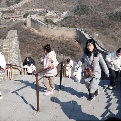 Ein Blick von oben auf einen steilen Treppenabschnitt der Chinesischen Mauer. Im Vordergrund steigen mehrere Personen, darunter eine Frau in einem langen grauen Mantel, die Stufen hinauf. Die massive Steinmauer schl&auml;ngelt sich im Hintergrund &uuml;ber karge, br&auml;unliche Gebirgsk&auml;mme bis zu den fernen Berggipfeln unter einem klaren blauen Himmel.