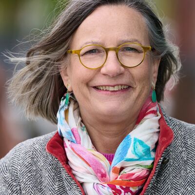 Nahaufnahme von Annemarie Stoltenberg, die freundlich in die Kamera l&auml;chelt. Sie hat graumeliertes, kurzes Haar, tr&auml;gt eine markante gelbe Brille und gr&uuml;ne Ohrh&auml;nger. Ihr Outfit besteht aus einer grau-wei&szlig; gemusterten Jacke mit Fischgr&auml;tmuster und einem farbenfrohen, gemusterten Schal in Blau-, Rosa- und Rott&ouml;nen. Der Hintergrund ist unscharf und zeigt nat&uuml;rliche Gr&uuml;n- und Braunt&ouml;ne.