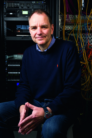 Bild vergr&ouml;&szlig;ern: Portr&auml;t eines Mannes in einem IT-Serverraum. Er sitzt mit ineinandergelegten H&auml;nden im Vordergrund und blickt l&auml;chelnd in die Kamera. Er tr&auml;gt einen dunkelblauen Pullover &uuml;ber einem hellblauen Hemd und Jeans. Im Hintergrund sind Serverschr&auml;nke mit technischem Equipment und ein B&uuml;ndel bunter Netzwerkkabel zu sehen.