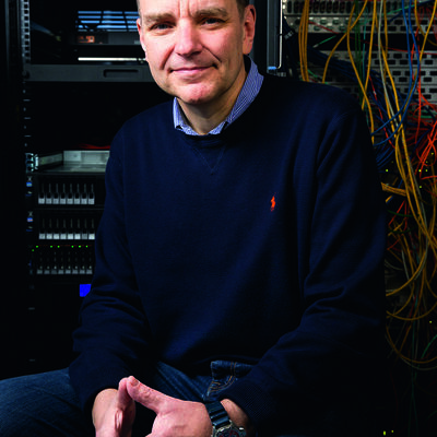Portr&auml;t eines Mannes in einem IT-Serverraum. Er sitzt mit ineinandergelegten H&auml;nden im Vordergrund und blickt l&auml;chelnd in die Kamera. Er tr&auml;gt einen dunkelblauen Pullover &uuml;ber einem hellblauen Hemd und Jeans. Im Hintergrund sind Serverschr&auml;nke mit technischem Equipment und ein B&uuml;ndel bunter Netzwerkkabel zu sehen.