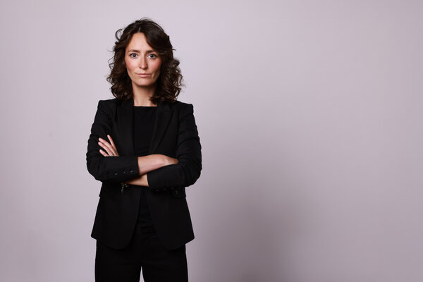 Bild vergr&ouml;&szlig;ern: Portr&auml;t einer Frau mit dunklen, schulterlangen Locken vor einem neutralen, hellgrauen Hintergrund. Sie tr&auml;gt einen schwarzen Blazer &uuml;ber einem schwarzen Oberteil und blickt mit verschr&auml;nkten Armen und einem direkten, seri&ouml;sen Ausdruck in die Kamera.