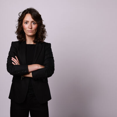 Portr&auml;t einer Frau mit dunklen, schulterlangen Locken vor einem neutralen, hellgrauen Hintergrund. Sie tr&auml;gt einen schwarzen Blazer &uuml;ber einem schwarzen Oberteil und blickt mit verschr&auml;nkten Armen und einem direkten, seri&ouml;sen Ausdruck in die Kamera.