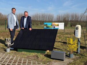 Zwei M&auml;nner in Sakkos stehen im Freien neben einem gro&szlig;en Solarmodul und einer E-Ladestation der Stadtwerke Geesthacht. Im Hintergrund ist ein Banner f&uuml;r das ,Natur- und Gartenfest' am 19.04.2026 zu sehen. Die Szene wird durch zwei bl&uuml;hende Topfpflanzen und einen blauen Himmel erg&auml;nzt.