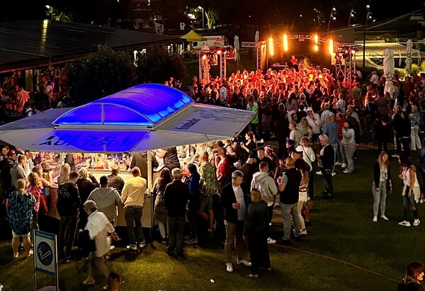 Bild vergr&ouml;&szlig;ern: Eine belebte Open-Air-Veranstaltung bei Nacht. Im Vordergrund steht ein gro&szlig;er Getr&auml;nkestand mit blau leuchtendem Dach, um den sich viele G&auml;ste versammeln. Im Hintergrund ist eine hell erleuchtete B&uuml;hne mit einer tanzenden Menschenmenge zu sehen. Die Atmosph&auml;re ist festlich und voller Energie.