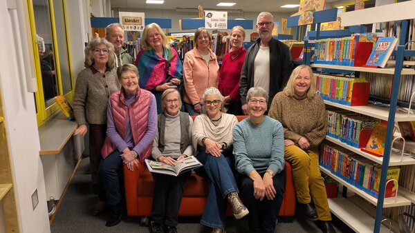 Eine Gruppe von elf l&auml;chelnden Erwachsenen - neun Frauen und zwei M&auml;nner - posiert f&uuml;r ein Gemeinschaftsfoto in einer gem&uuml;tlichen Bibliothek. Vier Personen sitzen auf einem orangefarbenen Sofa im Vordergrund, eine von ihnen h&auml;lt ein aufgeschlagenes Buch. Sieben weitere Personen stehen dahinter. Die Gruppe ist umgeben von gut gef&uuml;llten B&uuml;cherregalen mit Kinder- und Jugendb&uuml;chern; im Hintergrund sind Schilder mit der Aufschrift &raquo;Klassiker&laquo; und &raquo;Science Fiction&laquo; zu sehen.