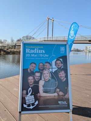 Bild vergr&ouml;&szlig;ern: Ein blaues Werbeplakat steht in einem silbernen Kundenstopper im Freien auf einer h&ouml;lzernen Uferpromenade. Im Hintergrund sind die Elbe, eine moderne Fu&szlig;g&auml;ngerbr&uuml;cke (Hafenbr&uuml;cke Geesthacht) und ein strahlend blauer Himmel zu sehen.
Details auf dem Plakat: Event: Musik am Hafen Geesthacht.
K&uuml;nstler: Die Band &raquo;Radius&laquo; (ein Portr&auml;tfoto zeigt sieben Bandmitglieder, eine Frau im Vordergrund und sechs M&auml;nner dahinter).
Datum & Uhrzeit: 20. Mai, 19 Uhr.
Ort: Sitzterrassen / Menzer-Werft-Platz.
Logos: Unten links ein Anker-Logo mit dem Text &raquo;Musik am Hafen Geesthacht&laquo;, unten rechts das &raquo;Moin Geesthacht&laquo;-Logo.
Besonderheit: Eine blaue Beachflag mit dem Veranstaltungslogo weht rechts hinter dem Aufsteller.