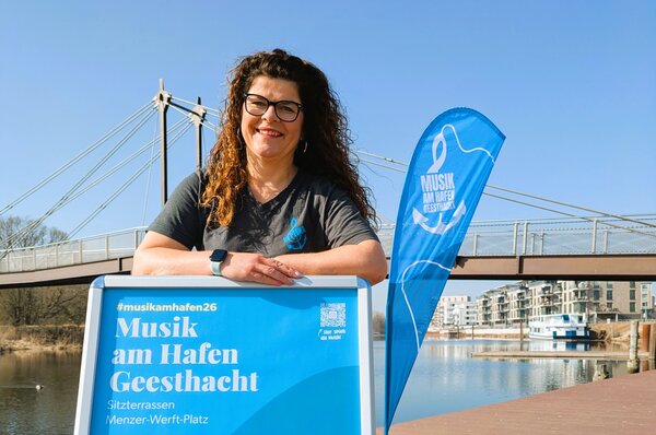 Eine l&auml;chelnde Frau mit braunem, lockigem Haar und Brille steht an der Hafenpromenade in Geesthacht. Sie lehnt sich entspannt auf einen silbernen Plakatst&auml;nder. Sie tr&auml;gt ein dunkelgraues T-Shirt und eine Smartwatch.
Hintergrund und Details:
Das blaue Plakat im St&auml;nder zeigt den Schriftzug &raquo;Musik am Hafen Geesthacht&laquo; sowie den Ort &raquo;Sitzterrassen / Menzer-Werft-Platz&laquo;.
Im Hintergrund erstreckt sich die Elbe unter einem strahlend blauen Himmel.
Die moderne Hafenbr&uuml;cke und eine blaue Beachflag mit dem Veranstaltungslogo sind deutlich zu sehen.